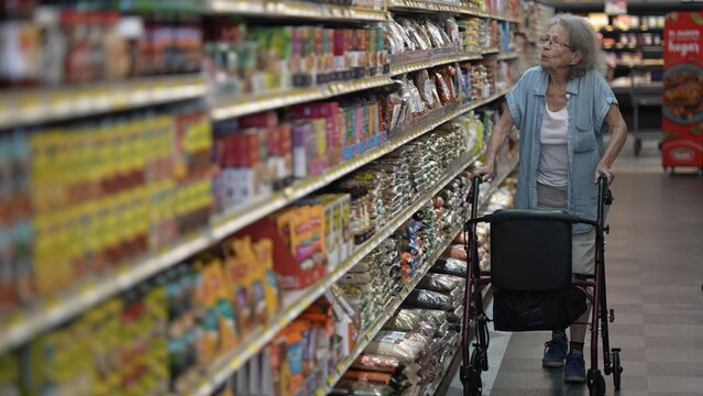 An elderly woman navigates a grocery store aisle with her walker, carefully selecting items from the shelves during her shopping trip in the supermarket.
