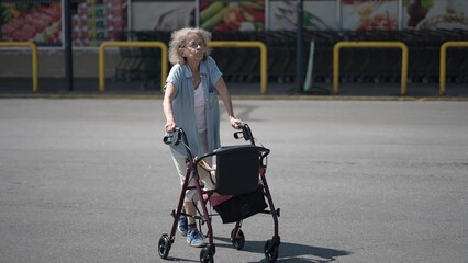 Obraz premium An elderly woman with silver hair relies on her walker as she navigates a grocery store parking lot, preparing for her shopping trip in broad daylight.