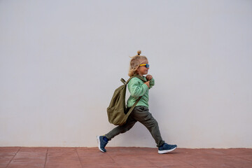 Happy young boy with blond hair in a ponytail, wearing sunglasses, green sweatshirt, and backpack,...