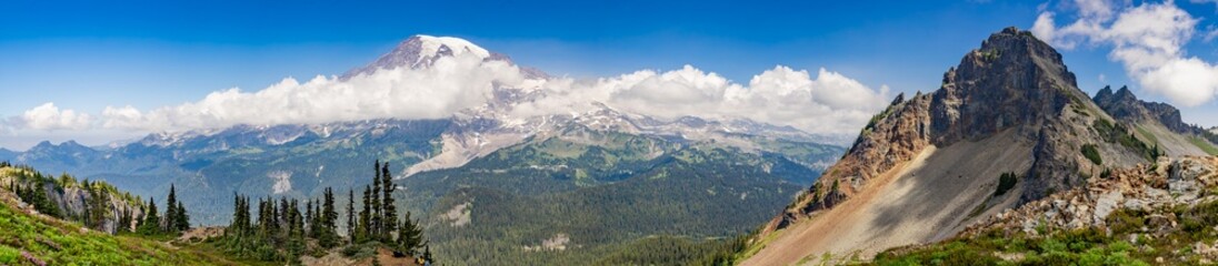 Fototapeta premium Stunning panoramic mountain view with a snow-capped peak, rocky ridges, evergreen forests, and drifting clouds. Ideal for themes of nature,travel, hiking, adventure, wilderness,and outdoor exploration