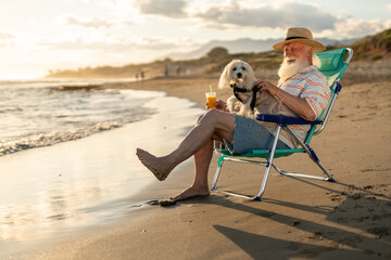 Happy senior man with white beard relaxing in a beach chair with his small dog on his lap, holding orange juice, enjoying sunset, pet companionship and peaceful vacation by the sea.