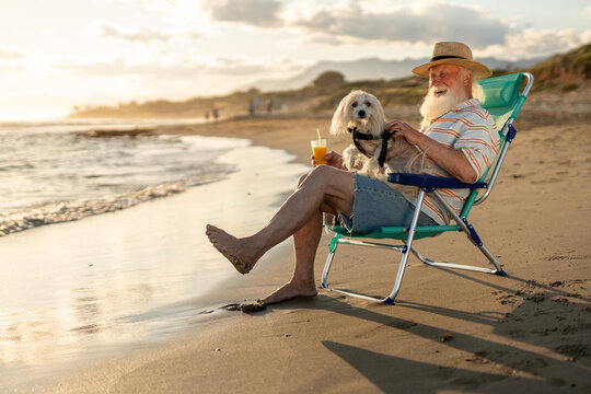 Happy senior man with white beard relaxing in a beach chair with his small dog on his lap, holding orange juice, enjoying sunset, pet companionship and peaceful vacation by the sea.