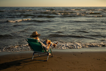 Elderly man in straw hat sitting alone in beach chair, enjoying orange juice and ocean waves during golden hour, reflecting peacefully while looking out at the endless sea horizon.