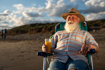 Elderly man with long white beard sitting on beach chair, wearing striped shirt and straw hat,...