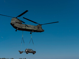 Military helicopter transporting ground vehicle via external sling load against clear sky