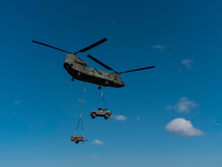Military helicopter carrying two vehicles simultaneously using sling load system under blue sky