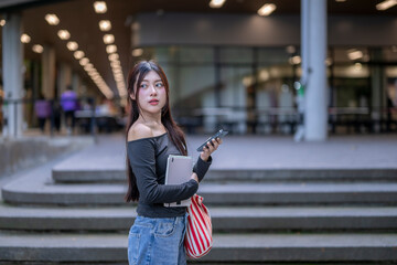 Fototapeta premium University student using smartphone and holding book outside college building