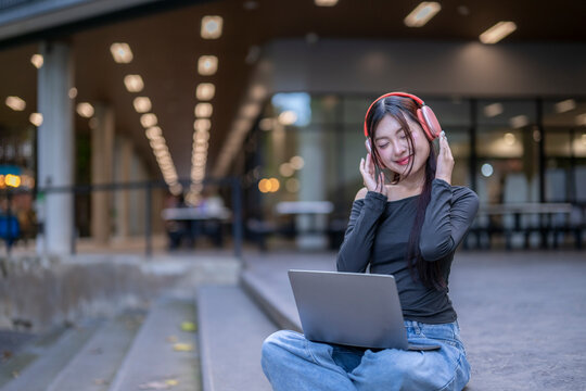 Young asian woman listening to music on laptop with headphones outdoors