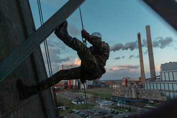 Soldier rappelling down industrial structure with power plant smokestacks visible in evening sky