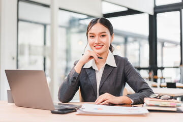 Confident businesswoman in gray suit sitting at modern office desk with laptop, documents, and...