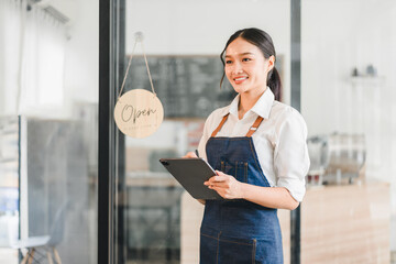 Young Asian woman barista wearing apron holding digital tablet standing in modern coffee shop with open sign, smiling and ready to serve customers