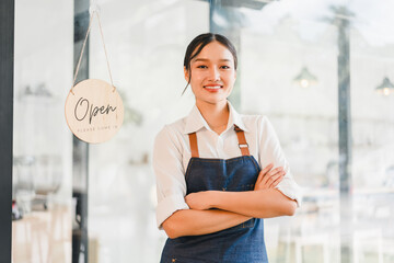 Young Asian woman barista wearing apron standing with arms crossed in front of coffee shop entrance, smiling confidently, open sign hanging on glass door, modern cafe interior visible inside