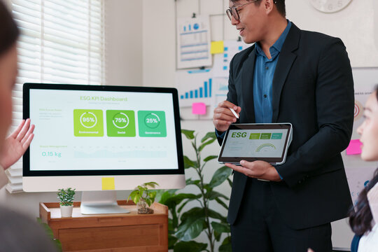 Businessman presenting ESG KPI dashboard on tablet and computer screen in modern office, discussing sustainability performance with colleagues, teamwork and data analysis concept