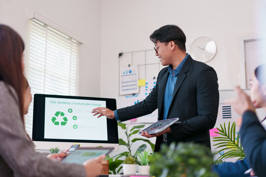 Businessman presenting ESG strategy with recycling symbol on screen, diverse team meeting in modern office, discussing sustainable future, teamwork, green plants, technology, positive atmosphere