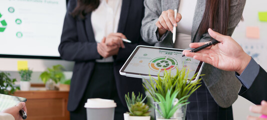 Business meeting with professionals discussing sustainability data on digital tablet, surrounded by...