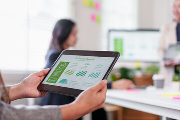 Person holding tablet with business charts, showing ESG impact overview in bright office environment with blurred colleagues in background, conveying productivity and teamwork