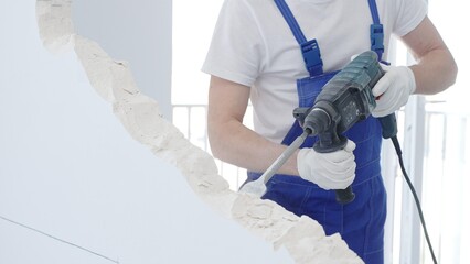 Male construction worker demolishing white wall with rotary hammer drill, wearing blue overalls and...