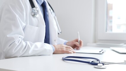 Blue stethoscope is lying on the table near doctor man professional writing on the clipboard in clinic office. Medicine and healthcare concept
