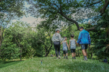 Fototapeta premium A happy Asian family with two young sons enjoys a walk in the park. Parents hold their children's hands while exploring a lush green hill together, a concept of family bonding.
