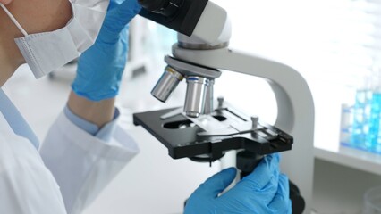 Close up of scientific professional blue gloved hands examining microscope lens, performing...