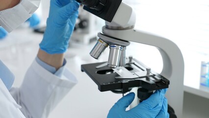 Close up of scientific professional blue gloved hands examining microscope lens, performing...