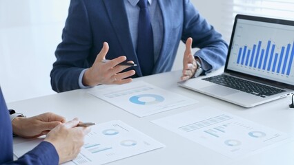 Business team analyzing financial data on a laptop and printed documents, pointing at charts and graphs during a meeting. Business people and finance concept