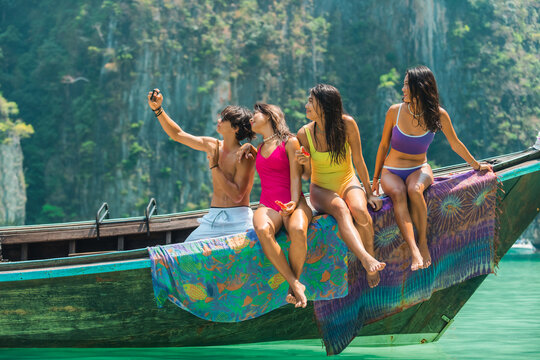 Happy Asian man and woman friends using mobile phone taking selfie together while travel on boat at tropical island in sunny day. People enjoy outdoor lifestyle on summer beach holiday vacation.
