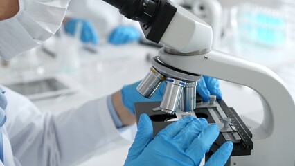 Close up of scientific professional blue gloved hands examining microscope lens, performing...