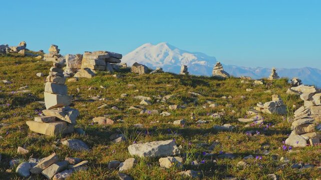 4k, panoramic view of the Bolshoy Bermamyt plateau with stone turrets built on a grassy slope, with the snow-capped peaks of Mount Elbrus in the background, at early morning, Stavropol Krai, Russia