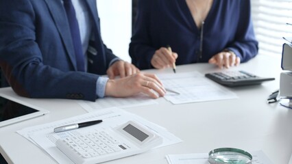 Close up of calculator and magnifying glass over financial documents opposite financial team analyzing data, reviewing reports, collaborating at modern office workspace. Audit and taxes in business