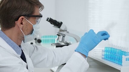 Masked male scientist in lab coat, protective glasses and blue gloves is adding reagent to test...