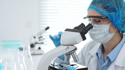 Female scientist analyzing sample through microscope, wearing protective lab gear during medical...