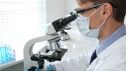 Masked scientist analyzing sample under microscope, colleague researching nearby in sterile medical...