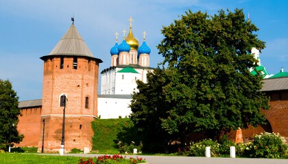 Obraz premium A picturesque scene of a historic red brick tower and a traditional Russian church, with lush green trees and a clear blue sky.