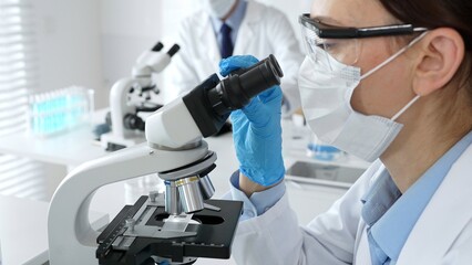 A female scientist in a blue mask and gloves is using a microscope and conducting scientific...