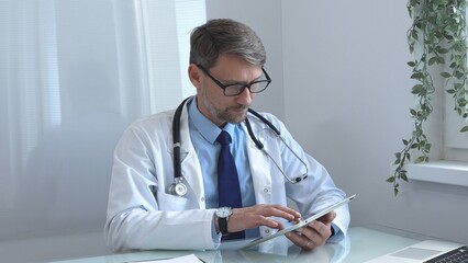 Medical doctor wearing lab coat and stethoscope using digital tablet consulting patient data in...