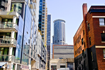 Fototapeta premium A narrow urban alley framed by brick and glass facades, leading to sleek high-rises and a round tower, blending industrial charm with modern city architecture.