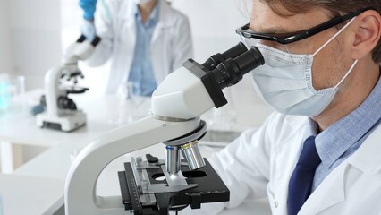 Masked scientist analyzing sample under microscope, colleague researching nearby in sterile medical...