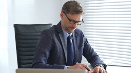 Adult businessman wearing blue suit, tail and glasses is concentrating while writing on a document...