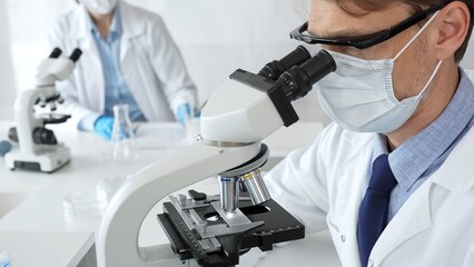 Masked scientist analyzing sample under microscope, colleague researching nearby in sterile medical...