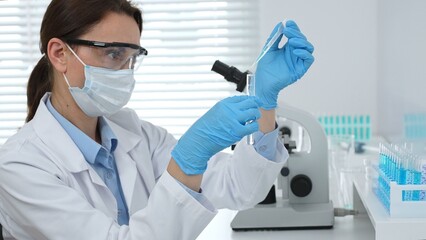 Female scientist wearing a face mask and blue gloves, carefully dropping liquid from a pipette into...