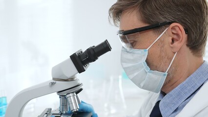 Scientist wearing mask and gloves using microscope doing scientific research in a laboratory with...