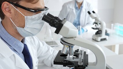 Medical researcher analyzing sample under microscope, wearing protective gear in sterile laboratory...