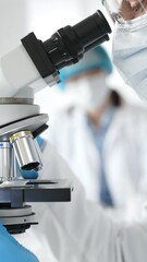 Close up of male researcher examining sample through microscope, wearing blue safety gear in modern laboratory setting. Medicine, healthcare and science concept