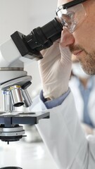 Close up of male researcher examining sample through microscope, wearing safety eyewear in sterile laboratory setting, surrounded by scientific glassware. Medicine, healthcare and science concept