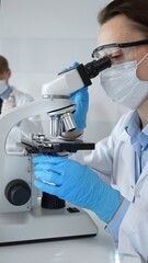 Female scientist wearing mask and blue gloves using microscope doing scientific research in modern laboratory with colleague working in background. Medicine, healthcare and science concept