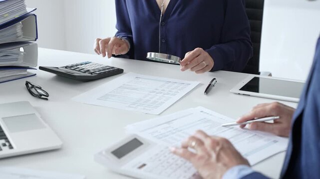 Female financial analysts examining documents, using magnifying glass and calculator while studying corporate data at workplace with a male colleague. Audit and taxes in business