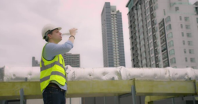 A engineer construction worker wearing a reflective safety vest and hard hat drinks water from a plastic bottle while standing on a building site with modern skyscrapers in the background