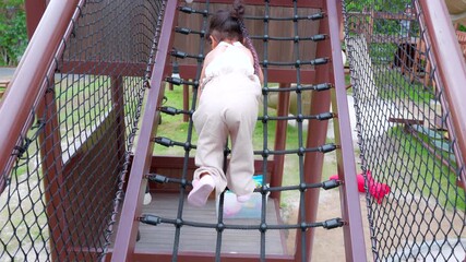 Happy Young Asian child girl climbing a rope bridge at a playground, showcasing outdoor play and adventure. - Powered by Adobe