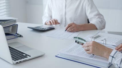 Two accountants are working together, using a magnifying glass and a calculator, to audit financial...
